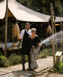 Webdesign Limburg | Two people celebrate outdoors, spraying champagne on a wooden boardwalk with a cream tent behind them.