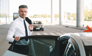 Webdesign Limburg | Chauffeur in a white shirt and black tie standing beside an open car door, jacket slung over his shoulder at a transport pickup.